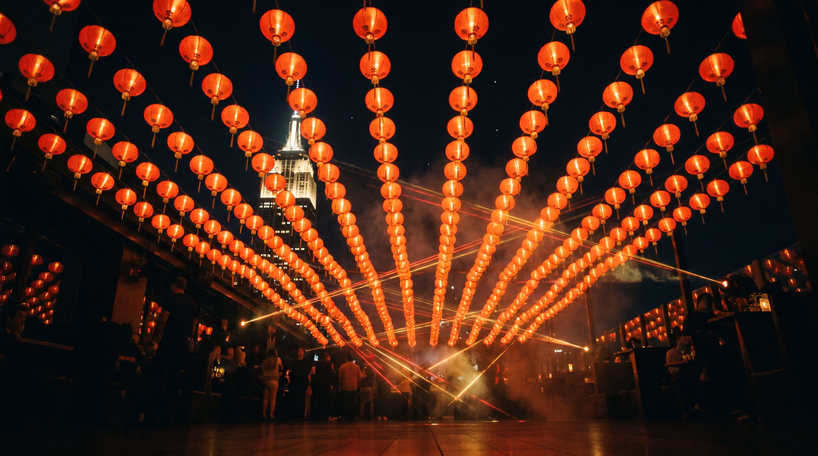 Looking up through a canopy of glowing red paper lanterns with red and gold lasers crossing above the rooftop