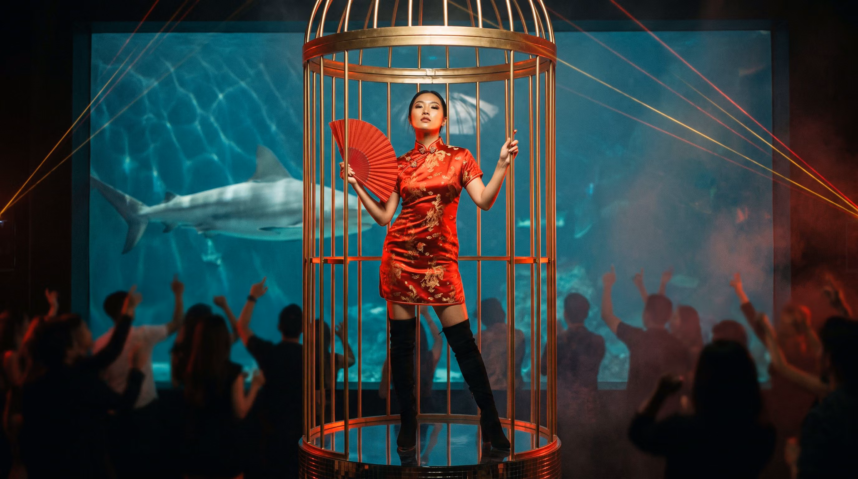A dancer in a red silk qipao with a red fan inside a gold cage in The Deep, a reef shark gliding behind the aquarium glass