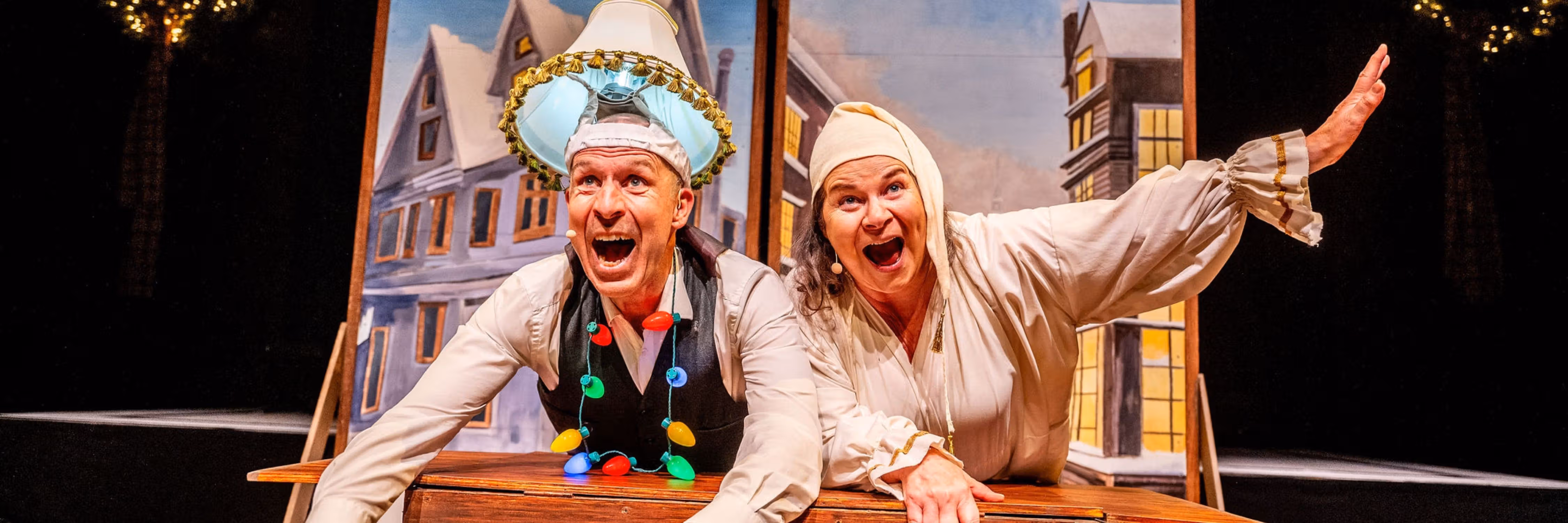 A man (Kerry) wearing a lamp shade on his head and Christmas lights around his neck rides a desk next to a woman (Linda) in nightwear from the 1800s.