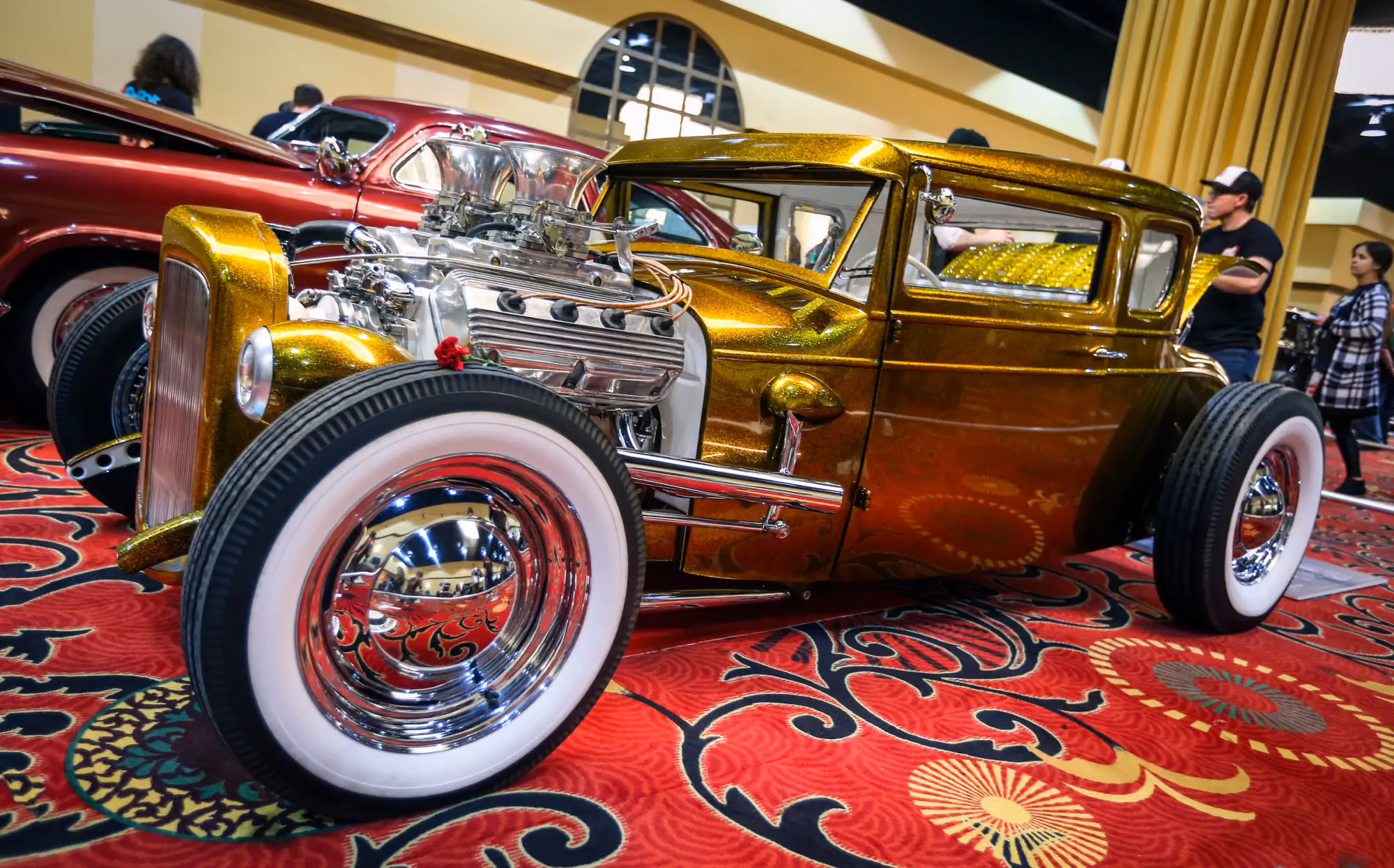A gold Model A hot rod with whitewall tires on the red show carpet