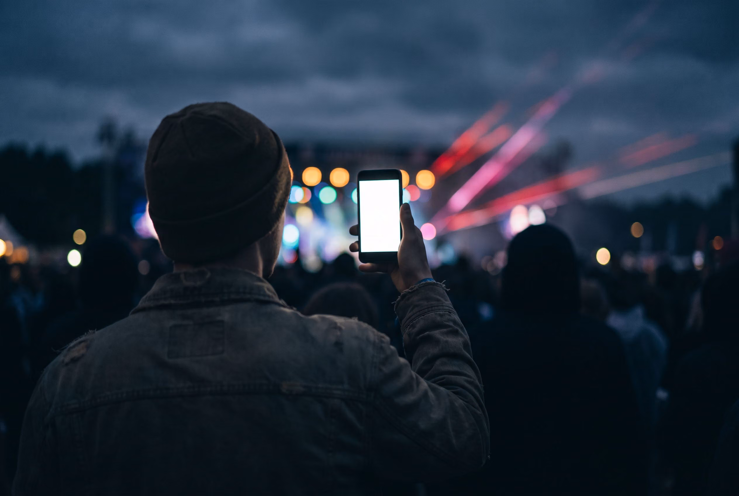 Festivalgoer holding up a phone in a crowd at night with stage lights behind