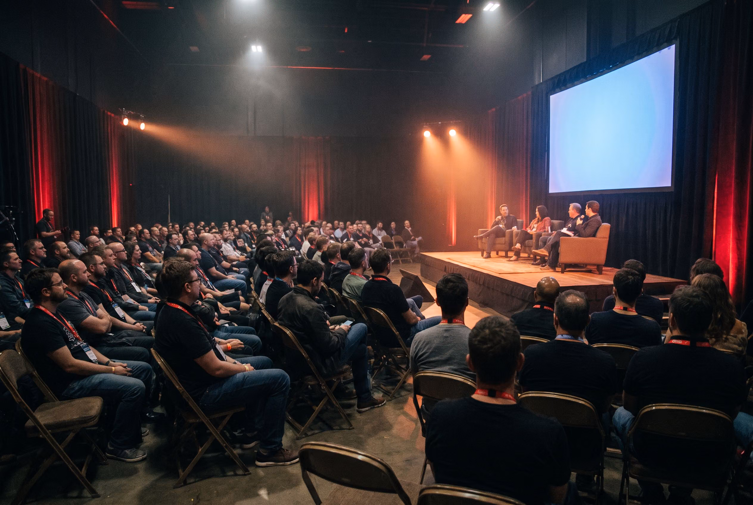 A full convention panel room with a seated audience facing four panelists on stage