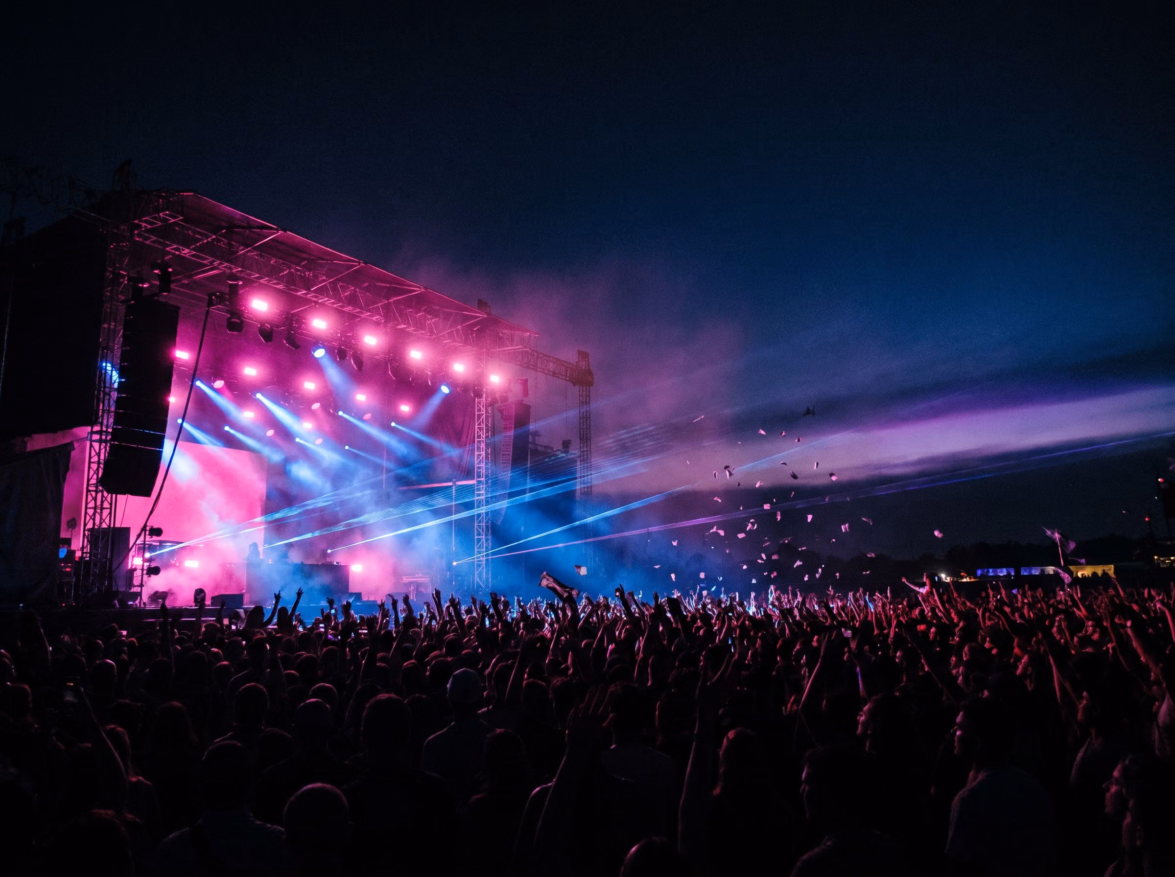 Festival crowd facing a main stage lit pink and blue at dusk